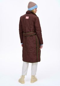 Br�ązowy pikowany długi płaszcz z zielonymi lamówkami, przewiązywany paskiem w talii i różową metką z tyłu. Stylizowany z jasnoniebieską czapką.