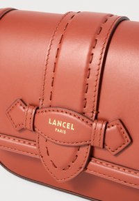 Borsa a mano in pelle marrone con un design strutturato, caratterizzata da accenti cuciti, un dettaglio a fiocco e il marchio impresso "LANCEL PARIS" in oro.