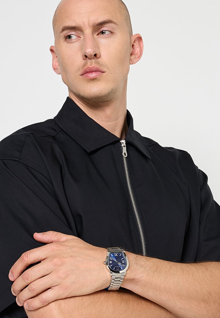 Silberne Uhr mit blauer Ziffernblatt, ausgestattet mit silbernen Stundenmarkierungen und Zeigern. Getragen an einem Handgelenk mit heller Haut, kombiniert mit einer schwarzen Jacke mit Kragen.