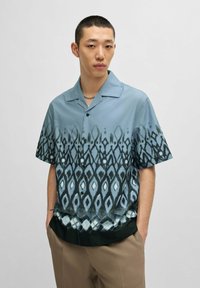 Camicetta a maniche corte di colore azzurro chiaro con un intricato motivo scuro che presenta diamanti e linee, realizzata in un tessuto morbido con una superficie liscia.