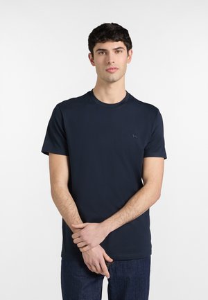 Mladý muž s krátkymi tmavými vlasmi, oblečený v obyčajnom tmavomodrom tričku a tmavých džínsach, stojaci so založenými rukami pred sebou.