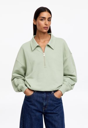 Frau mit langen dunklen Haaren, die einen hellgrünen Sweatshirt mit Reißverschluss und blaue Jeans trägt, steht mit Händen in den Taschen vor weißem Hintergrund.