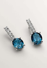 Pendientes de plata con grandes piedras azules ovaladas facetadas, acompañadas de pequeñas piedras transparentes a lo largo del pasador. Montura de metal texturizado.