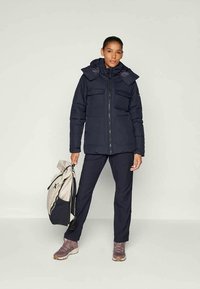 Mujer con una chaqueta de invierno negra con capucha y pantalones, sosteniendo una mochila beige y negra, de pie frente a un fondo claro y uniforme.