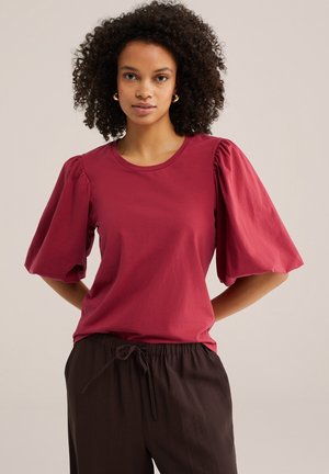 Femeie cu păr creț purtând cercei mari aurii, un top roșu cu mâneci bufante și pantaloni închiși la culoare cu șnur, pozând cu mâinile la spate.