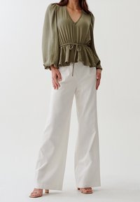 Blusa verde oliva, con texture e scollo a V, vita stretta e orlo sbuffato, abbinata a pantaloni bianchi a gamba larga e sandali rosa chiaro.