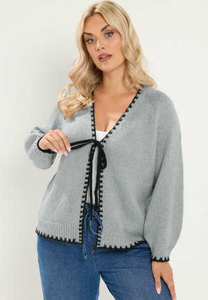 Blonde vrouw die een lichtgrijze gebreide cardigan met zwarte bies draagt en een zwart lint aan de voorkant vastknoopt, gecombineerd met een blauwe spijkerbroek en een wit topje.