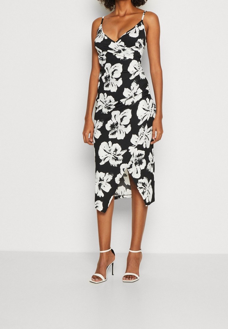 Žena v černých midi šatech s bílým květinovým potiskem, s tenkými ramínky, předním rozparkem a bílými sandály na vysokém podpatku.