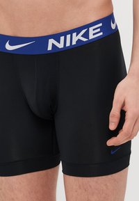 Fekete sportos boxeralsó kék elasztikus derékpánttal, rajta fehér "NIKE" logóval. Sima textúra, testhezálló dizájn, minimális varrásokkal.