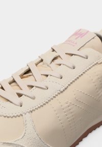 Μπεζ αθλητικό παπούτσι με πάνελ από σουέντ και λείο υλικό, μπεζ κορδόνια και ροζ ετικέτα με λογότυπο στη γλώσσα, εμφανιζόμενο σε κοντινή λεπτομέρεια.