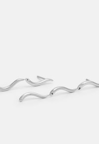 Brincos prateados em forma de onda com superfícies lisas e refletivas e elos interconectados, apresentando um design minimalista.