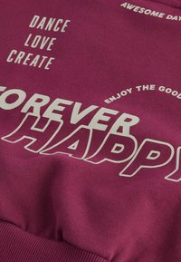 Fioletowy sweatshirt z białym napisem; zawiera takie frazy jak "NA ZAWSZE SZCZĘŚLIWY" i "TAŃCZ KOCHAJ TWÓRZ". Miękka, przypominająca bawełnę tekstura, ściągacze przy mankietach.