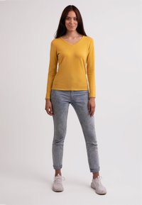 Gul långärmad V-ringad topp i kombination med ljusblå jeans och grå sneakers. Slät yta, figurnära passform, uppvikt nederkant på byxorna.