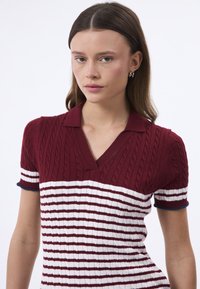 Polo en tricot avec des rayures horizontales rouge foncé et blanches. Design à col en V avec un côtelé texturé et des accents en azul marine contrastants sur les manches.