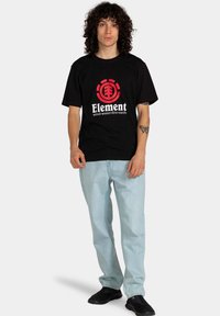 Camiseta de algodón negra con un logo circular rojo que contiene el texto "Element" y el eslogan "viento-agua-fuego-tierra"; combinada con jeans de color azul claro y zapatos negros.