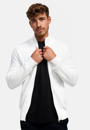 Weiße strukturierte Zip-Jacke mit Stehkragen, gerippten Bündchen und Saum. Tragen über einem schwarzen Shirt, mit beiden Händen am Kragen gehalten.
