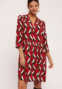 Czerwona i czarna sukienka w wzór z dekoltem w kształcie litery V, rękawami długości 3/4, z talią ściągniętą oraz peplum na dole, zdobiona geometrycznymi kształtami w kolorze kremowym i brązowym.