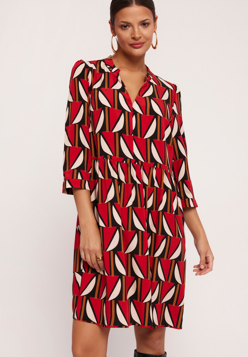 Czerwona i czarna sukienka w wzór z dekoltem w kształcie litery V, rękawami długości 3/4, z talią ściągniętą oraz peplum na dole, zdobiona geometrycznymi kształtami w kolorze kremowym i brązowym.