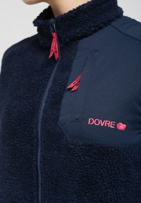 Marineblå fleecejakke med en glat stofpanel, høj krave, røde lynlåsdragere og broderet "DOVRE" logo på venstre bryst.