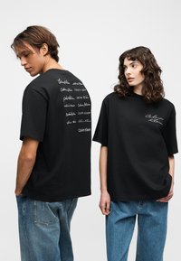 Un homme et une femme portant des T-shirts noirs Calvin Klein avec le texte du logo ; l'homme montre le dos du T-shirt, la femme montre le logo à l'avant, tous deux en jeans bleus.