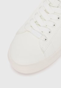Sneaker sintetica bianca con una texture liscia, punta rotonda e lacci bianchi piatti. Presenta un sottile logo "GUESS" sul lato.