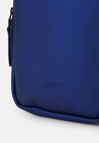Zaino impermeabile blu realizzato in materiale sintetico liscio. Presenta il logo "RAINS" in rilievo nella parte inferiore e un design elegante e minimalista.