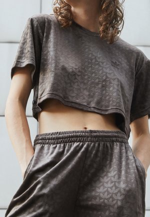 Osoba ubrana w dopasowany, teksturowany ciemnoszary krótki top i spodnie Adidas, z rękami w kieszeniach, pokazana od talii do podbródka.