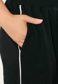 Svarta byxor med elastisk midja, med en vit sidestreck. En hand är placerad i en sidoficka, vilket framhäver tygets släta textur.