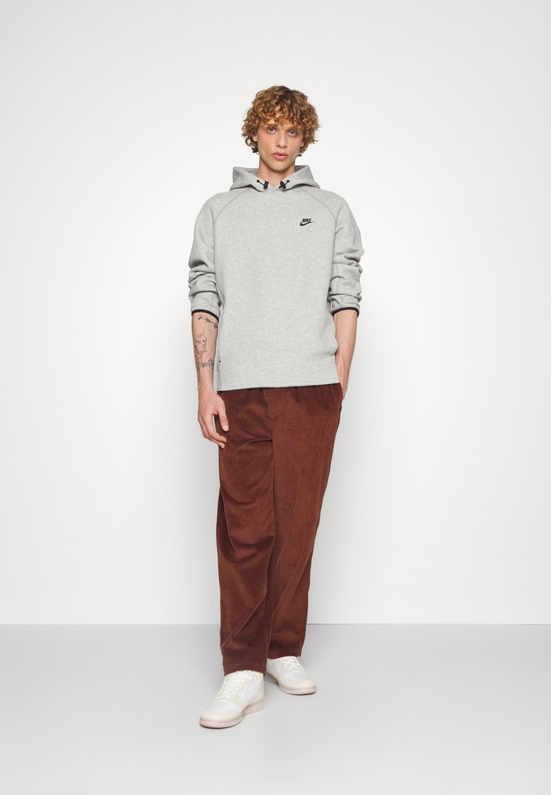 Haut à capuche gris avec logo noir, accents cordés ; pantalon en velours côtelé marron ; baskets blanches avec accents clairs. Coupe simple et décontractée, et matériaux texturés.