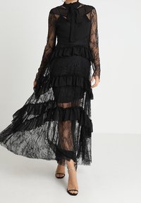 Abito in pizzo nero con maniche lunghe trasparenti, strati arricciati e un colletto con fiocco. Presenta un corpino aderente e una gonna fluida, che aggiungono texture ed eleganza.