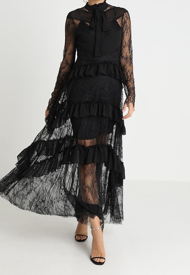 Abito in pizzo nero con maniche lunghe trasparenti, strati arricciati e un colletto con fiocco. Presenta un corpino aderente e una gonna fluida, che aggiungono texture ed eleganza.