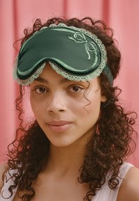 Groene satijnen slaapmasker met kantafwerking, voorzien van een geborduurde strikdetails aan de zijkant. Zachte textuur tegen een roze achtergrond.