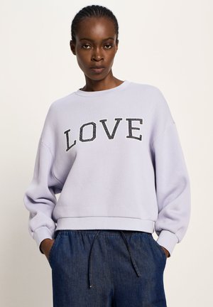 Woman with braided hair wearing a light purple sweatshirt with "LOVE" text and blue drawstring denim pants, hands in pockets, neutral background.