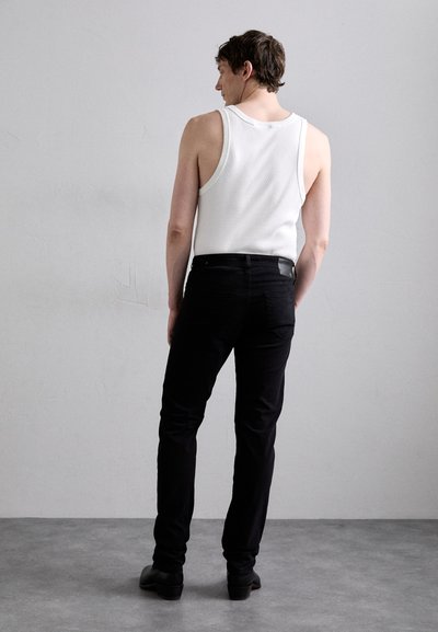 Λευκό αμάνικο μπλουζάκι και εφαρμοστά μαύρα τζιν με λεία υφή; μαύρα παπούτσια; στέκεται από πίσω σε ένα ανοιχτό γκρι φόντο.