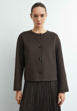 Femme portant une veste en laine marron foncé boutonnée et une jupe plissée assortie, debout devant un fond clair uni.