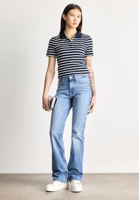 Fiatal nő hosszú, sötét hajjal, tengerészkék csíkos pólóingben, világoskék farmernadrágban, fehér tornacipőben, ezüst borítéktáska a kezében, áll.