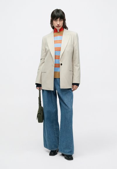 Μπεζ σακάκι, ριγέ πουλόβερ σε πορτοκαλί, μπλε και καφέ, τζιν με φαρδιά μπατζάκια, μαύρες μπότες, κρατώντας μια πράσινη τσάντα. Μινιμαλιστικός σχεδιασμός.