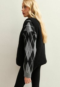 Svart fleeceväst med en strukturerad yta, som har grå ärmar med argyle-mönster och ribbade manschetter. Enkel, modern design.