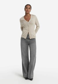 Beige, stickad kofta med V-ringning och framknappar, tillsammans med grå jeans med vida ben och svarta skor; slät textur och avslappnad passform.