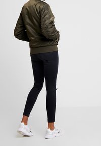 Giacca bomber verde oliva con polsini e orlo a costine, abbinata a jeans skinny neri con dettagli strappati e scarpe da ginnastica bianche.