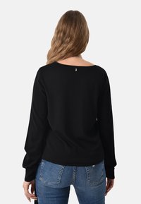 Maglione nero a maniche lunghe con scollo a goccia e polsini a coste. Presenta un piccolo dettaglio vicino al collo. Indossato con jeans in denim blu.