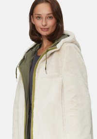 Manteau en fausse fourrure crème avec une doublure vert olive, fermeture éclair intégrale, capuche réglable avec cordon, et poches latérales. Texture douce avec une coupe décontractée.