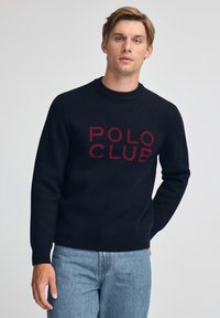 Junger Mann trägt einen schwarzen Pullover mit rotem "POLO CLUB" Schriftzug, kombiniert mit hellblauen Jeans, und steht vor einem schlichten hellgrauen Hintergrund.