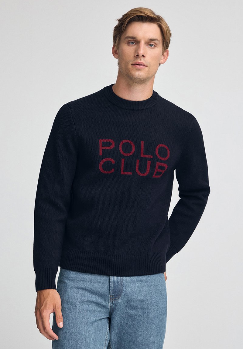 Junger Mann trägt einen schwarzen Pullover mit rotem "POLO CLUB" Schriftzug, kombiniert mit hellblauen Jeans, und steht vor einem schlichten hellgrauen Hintergrund.