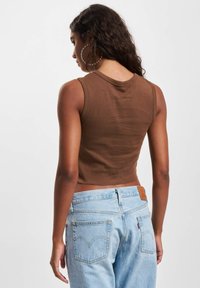 Brun avslappet kort topp uten ermer laget av mykt materiale, kombinert med lyseblå high-waisted denimjeans med baklommer og et lærmerke.
