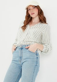 Chemise à rayures verte clair et blanche, associée à un jean en denim bleu taille haute. Le mannequin a les mains dans les poches, portant une casquette crème.