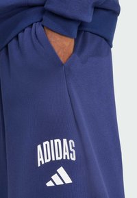 Tumši zils Adidas džemperis ar kabatu, kuram ir balts logs un trīs svītru dizains apakšējā kreisajā pusē. Mīksta auduma, ar brīvu piegriezumu.