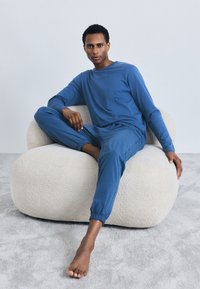 Blå bomulds loungewear sæt med lange ærmer og elastiske manchetter, båret mens man sidder på en cremefarvet, tekstureret, buet stol mod en lys baggrund.