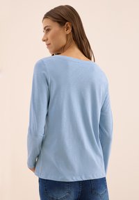 Chemise à manches longues bleu clair avec col arrondi et texture douce, assortie à un jean foncé. La vue de dos montre les coutures et un design simple.