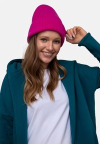 Turkusowa rozpinana bluza na białej koszulce, w połączeniu ze sztruksową, jasnoróżową czapką. Włosy są falujące i luźne, na neutralnym tle.